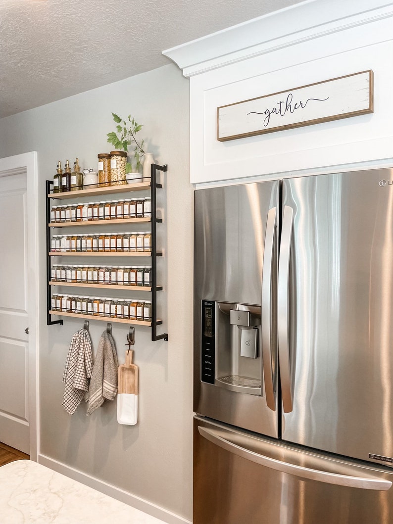 May include: A stainless steel refrigerator with a water dispenser and a wooden sign above it that reads "gather". A spice rack with many glass jars of spices is mounted on the wall to the left of the refrigerator.