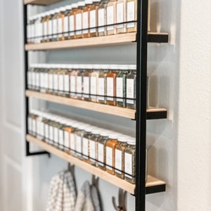 May include: A black metal spice rack with five wooden shelves filled with glass spice jars. The rack is mounted to a white wall and has hooks underneath for hanging towels.
