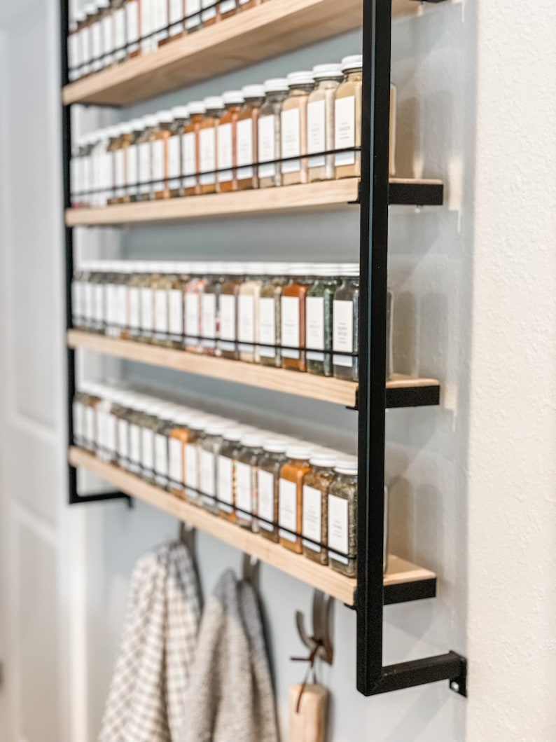 May include: A black metal spice rack with five wooden shelves filled with glass spice jars. The rack is mounted to a white wall and has hooks underneath for hanging towels.