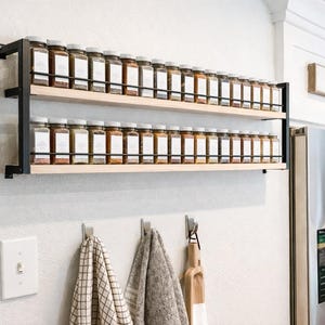 Peut inclure: Une étagère à épices à deux niveaux avec des supports en métal noir et des étagères en bois clair. Les étagères contiennent de nombreux pots d'épices en verre avec des étiquettes blanches. En dessous, des crochets maintiennent des torchons et un ustensile en bois.