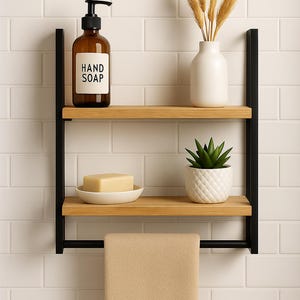 May include: A two-tier bathroom shelf with a black metal frame and light wood shelves. The top shelf holds a brown hand soap dispenser and a white vase with dried wheat. The bottom shelf has a small white potted plant and a bar of soap. A beige towel hangs below.