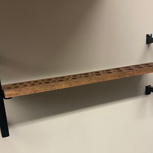 May include: A wooden shelf with a black metal frame. The shelf has a series of round holes drilled into the surface. The shelf is mounted to a wall.