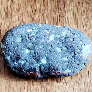 May include: A blue-grey, oval-shaped stone with a textured surface. The stone has patches of green, white, and reddish-brown hues. The stone is lying on a wooden surface.