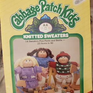 May include: A Cabbage Patch Kids knitting pattern booklet featuring three Cabbage Patch Kids dolls wearing knitted sweaters, pants, and skirts. The cover features the Cabbage Patch Kids logo and the text "Knitted Sweaters" and "14 Sweaters Plus Pants and Skirts - 23 Items in All".