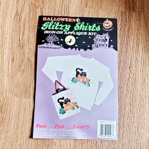 May include: A Halloween Glitzy Shirts iron-on applique kit featuring a white long-sleeve shirt and a tote bag decorated with a black cat and pumpkin graphic. The packaging includes the text Fast... Fun... Easy!!! and Kitty n' Pumpkin.