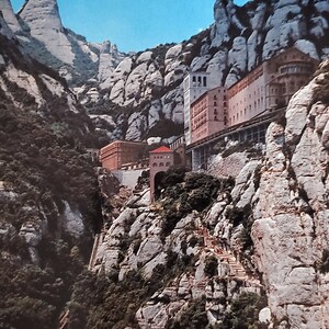 May include: A view of a monastery built into the side of a rocky mountain. The monastery is made of stone and has a red roof. There are trees and plants growing on the mountainside.