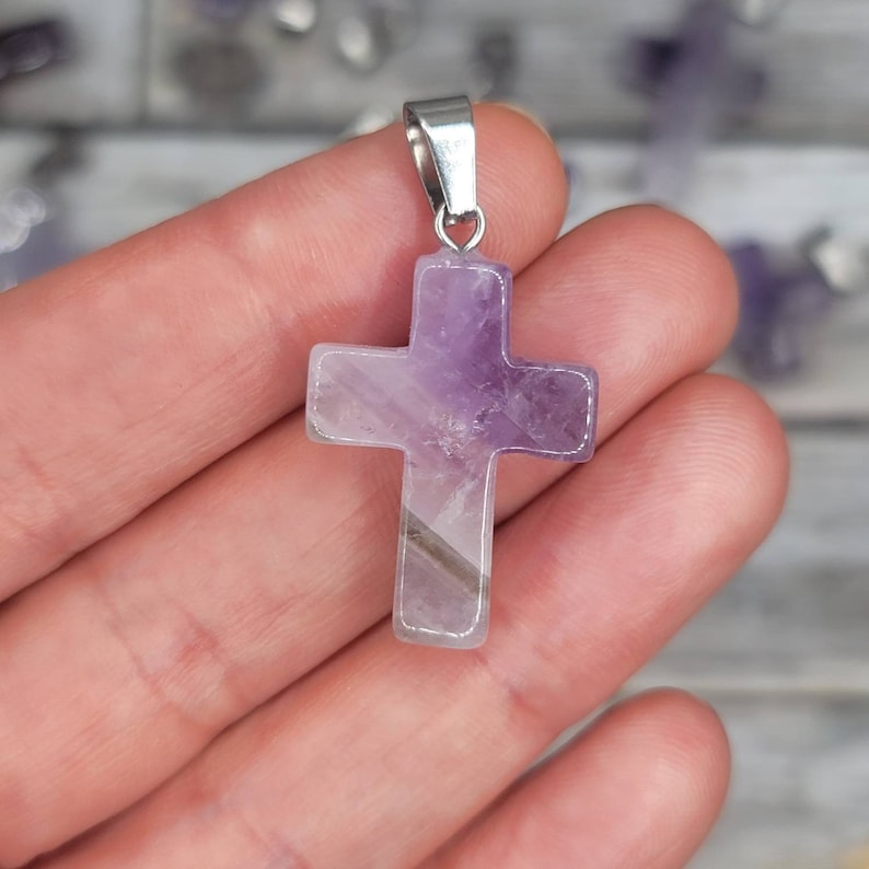May include: Amethyst cross pendant.  The cross is a deep purple color with lighter purple and grey streaks throughout. The cross is made of natural amethyst and has a polished, smooth finish. It features a simple, classic cross design with a slightly textured surface. The pendant hangs from a silver-toned metal bail. This gemstone cross is perfect for adding a touch of spiritual style to any outfit.