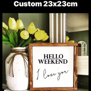 Op de afbeelding: Een houten bord met de tekst "Hello Weekend I love you" in zwarte letters op een witte achtergrond. Het bord is ingelijst in een donkerbruin houten frame en meet 23x23cm.