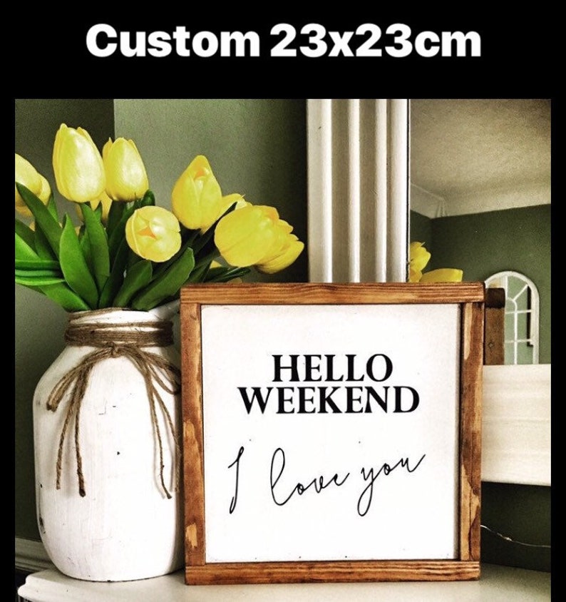 Op de afbeelding: Een houten bord met de tekst "Hello Weekend I love you" in zwarte letters op een witte achtergrond. Het bord is ingelijst in een donkerbruin houten frame en meet 23x23cm.