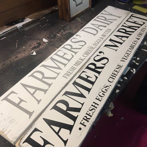 Peut inclure: Deux panneaux en bois blanc avec des lettres noires. Le panneau du haut indique "Farmers Dairy Fresh Milk, Cheese, Eggs & Butter". Le panneau du bas indique "Farmers &bull; Fresh Eggs, Cheese Vegetables & Fruits &bull;".