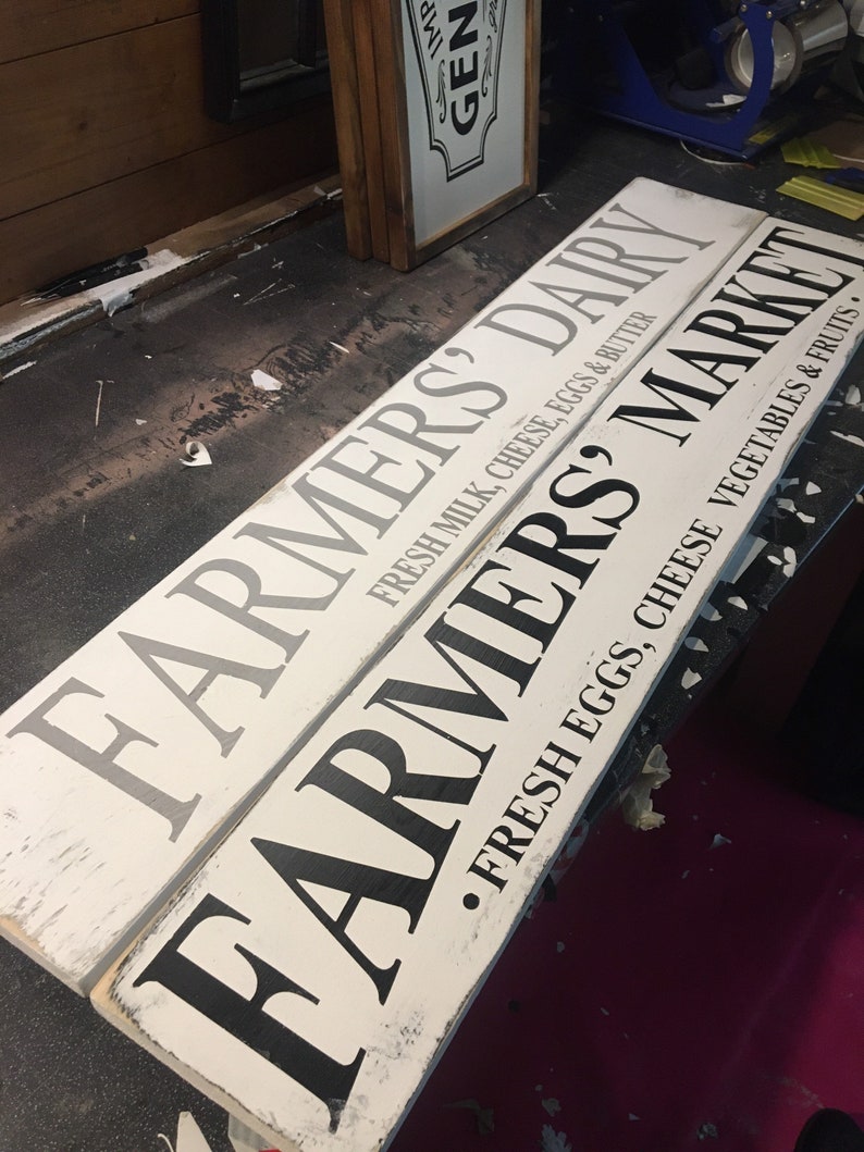 Peut inclure: Deux panneaux en bois blanc avec des lettres noires. Le panneau du haut indique "Farmers Dairy Fresh Milk, Cheese, Eggs & Butter". Le panneau du bas indique "Farmers &bull; Fresh Eggs, Cheese Vegetables & Fruits &bull;".