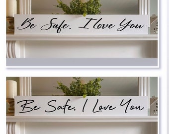 Op de afbeelding: Een wit houten bord met de woorden "Be Safe, I Love You" in zwart schrift. Het rechthoekige bord staat op een plank, met groen en kaarsen op de achtergrond. Een eenvoudige, oprechte boodschap.