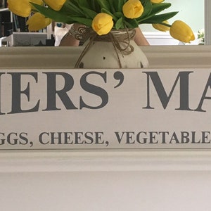 Peut inclure: Une enseigne en bois blanc avec le texte "FARMERS' MARKET" en lettres grises. Sous le texte, une ligne de texte plus petite indique "FRESH EGGS, CHEESE, VEGETABLES & FRUITS".