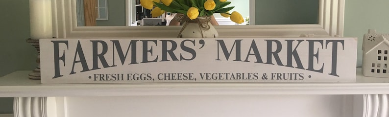 Peut inclure: Une enseigne en bois blanc avec le texte "FARMERS' MARKET" en lettres grises. Sous le texte, une ligne de texte plus petite indique "FRESH EGGS, CHEESE, VEGETABLES & FRUITS".