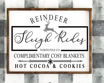 Op de afbeelding: Een wit houten bord met een bruin frame heeft de tekst "Reindeer Sleigh Rides" in zwarte cursieve letters met een rendier silhouet boven de tekst. Onder de tekst staat "North Pole Co. Complimentary Cosy Blankets Hot Cocoa & Cookies" in zwarte letters.
