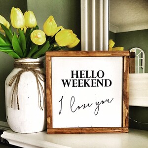Op de afbeelding: Een houten bord met de tekst "HELLO WEEKEND I love you" in zwarte letters. Het bord staat op een wit oppervlak met een vaas met gele tulpen op de achtergrond.
