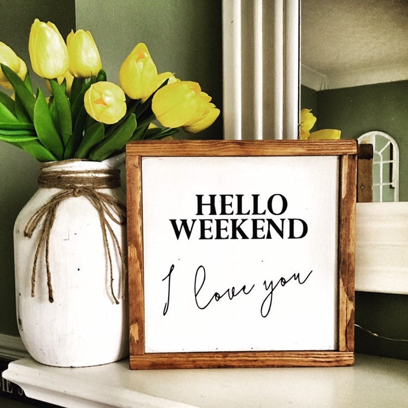 Op de afbeelding: Een houten bord met de tekst "HELLO WEEKEND I love you" in zwarte letters. Het bord staat op een wit oppervlak met een vaas met gele tulpen op de achtergrond.