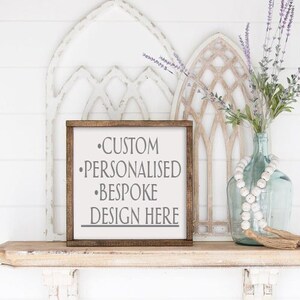 Op de afbeelding: Een houten bord met een witte achtergrond en een bruin frame. Het bord zegt "CUSTOM PERSONALISED BESPOKE DESIGN HERE" in grijze tekst. Het bord staat op een plank met een vaas met lavendel en een kralensnoer.