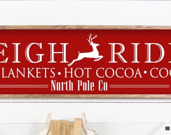 Op de afbeelding: Rood houten bord met witte tekst die "Sleigh Rides Warm Blankets Hot Cocoa Cookies North Pole Co." luidt. Een wit silhouet van een rendier springt tussen de woorden "Sleigh" en "Rides".