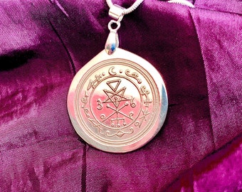 May include: A silver pendant with a circular design featuring a star, a cross, and other symbols. The pendant is hanging from a silver chain on a purple fabric background.