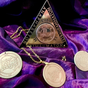 May include: Three gold-toned metal pendants with intricate designs and symbols. The pendants are on a purple fabric background. The pendant on the left has the text "EL TETRAGRAMMATON" and "PRIMEUMATON" on the triangle. The pendant on the right has the text "CHA NAPHAXETON MI NOLEION".