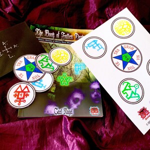 May include: A book titled "The Book of Fallen Darkness: Western Grimoire of Strong Magick" by Carl Nagel with a set of colorful stickers featuring various symbols and the text "Consecrated Talisman &copy;2019 Daemonic Dreams www.daemonicdreams.com"