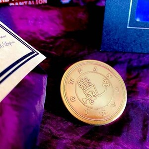 May include: A gold-colored metal coin with a circular design. The coin features a symbol with a cross and a hand holding a staff. The coin is surrounded by a circle of text that reads "N.D.A.N.I.D."