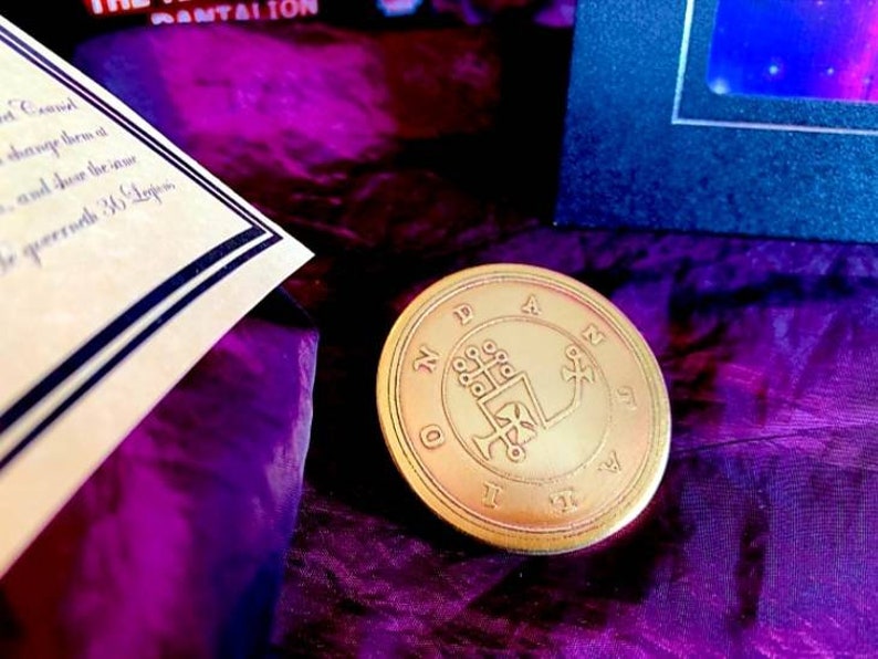May include: A gold-colored metal coin with a circular design. The coin features a symbol with a cross and a hand holding a staff. The coin is surrounded by a circle of text that reads "N.D.A.N.I.D."