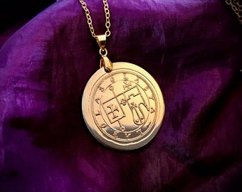 May include: A gold pendant necklace with a round, engraved charm featuring a complex geometric design. The charm has a circular border with letters and numbers around the edge.