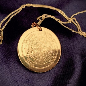 Könnte beinhalten: Eine goldene Halskette mit einem runden, gravierten Anhänger mit einem kreisförmigen Design mit Text. Der Anhänger befindet sich an einer goldenen Kette.