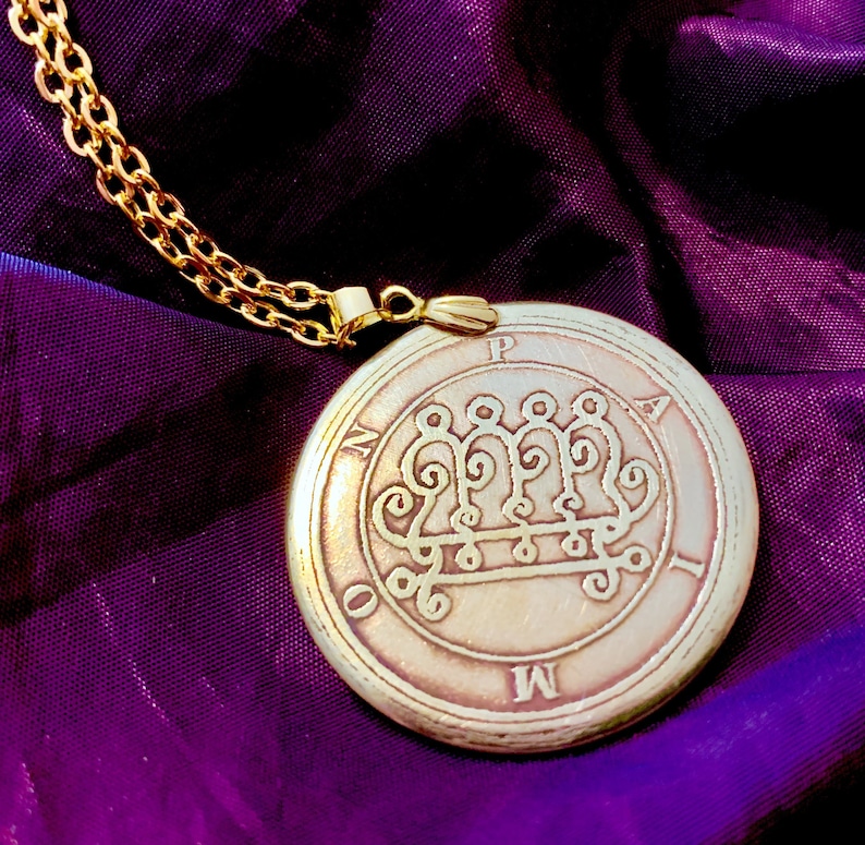 May include: A gold-colored pendant necklace with a circular, engraved pendant. The pendant features an intricate design with the letters "M", "O", "N", "P", and "A" around the edge. The necklace chain is also gold-colored, and the background is a deep purple.