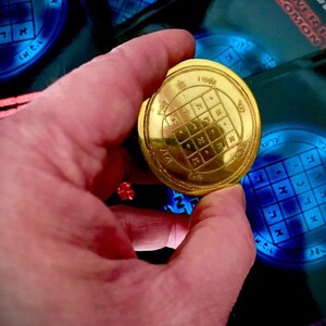 May include: A gold-colored metal disc with a square grid pattern and Hebrew lettering. The disc is being held in a hand.