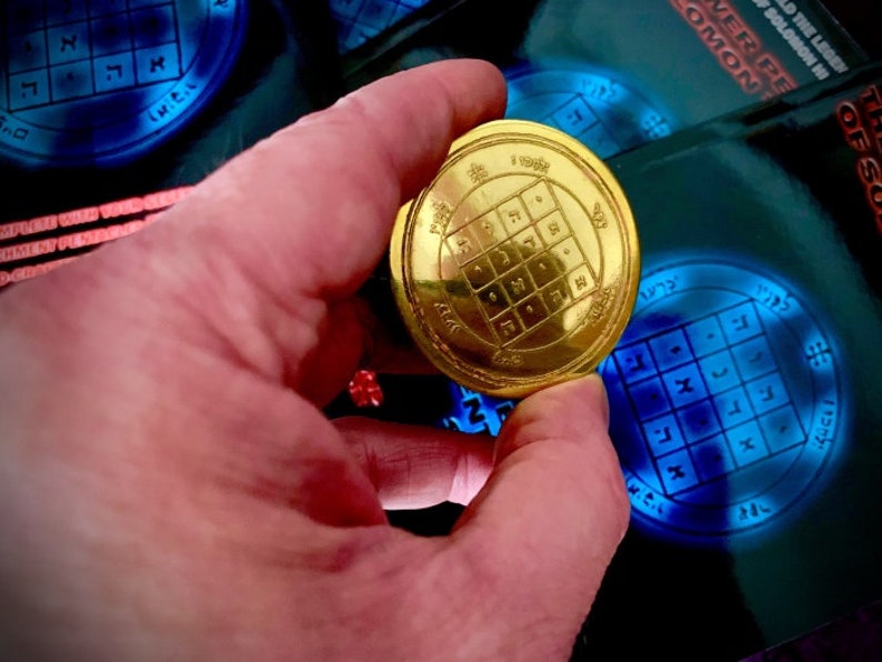 May include: A gold-colored metal disc with a square grid pattern and Hebrew lettering. The disc is being held in a hand.