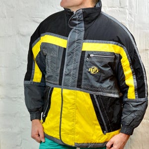 May include: A retro ski suit featuring a colour-blocked jacket and trousers. The jacket is black, grey, and yellow with a zip and pockets. The trousers are teal. The jacket has the text "RST" on the chest.