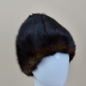 May include: A dark brown fur hat with a lighter brown trim. The hat is displayed on a white mannequin head. The fur appears soft and plush, with a slight sheen. The hat is a classic style, likely for cold weather.