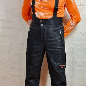 Puede incluir: Una camiseta de manga larga naranja con detalles blancos desgastados y la palabra DIESEL impresa en el pecho, combinada con pantalones de esquí negros de talle alto con tirantes negros ajustables, dos bolsillos laterales con cremallera y un logo GIERRE HTE.
