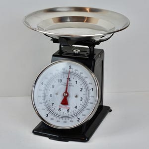 Op de afbeelding: Een vintage zwarte keukenweegschaal met een roestvrijstalen kom en een ronde wijzerplaat. De wijzerplaat heeft witte cijfers en een rode naald, en de weegschaal meet tot 2.27 kg.