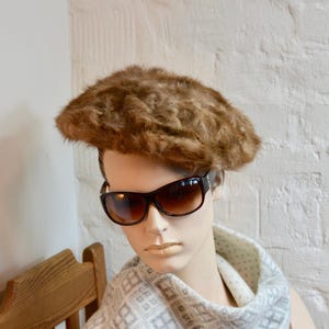 May include: A mannequin wearing a brown fur beret and dark brown sunglasses. The mannequin also has a light gray scarf with a geometric pattern. The background is a white brick wall.