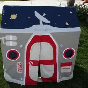 May include: A children's play tent designed as a space station. The tent features a gray and red exterior with a blue roof depicting a night sky with stars, a satellite dish, and the words "Space Station".
