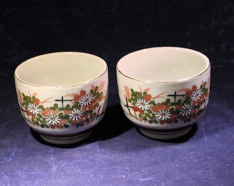 Puede incluir: Dos tazas de té de cerámica blanca con un diseño floral. Las tazas tienen un borde marrón y un interior blanco. El diseño floral presenta flores blancas, hojas verdes y detalles rojos.