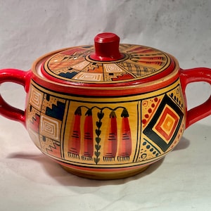 Può includere: Una zuccheriera in ceramica dipinta a mano con coperchio. La ciotola e il coperchio sono decorati con motivi geometrici rossi, gialli, neri e blu. Ha due manici rossi e un pomello rosso sul coperchio.