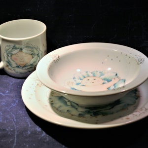 May include: A white ceramic mug, bowl, and plate with a blue and green floral design featuring a baby's face. The mug has a handle and the bowl and plate have a scalloped edge.