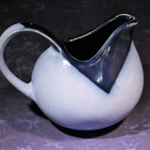 May include: A white ceramic creamer with a blue glaze. The creamer has a rounded body and a curved handle. The glaze is darker blue at the top of the creamer and fades to a lighter blue at the bottom.