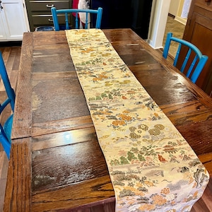 May include: A decorative table runner featuring a detailed pattern of landscapes, flowers, and foliage in gold, orange, green, and cream. The runner is placed on a dark wooden table, adding a touch of elegance.
