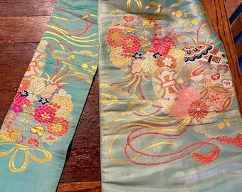 May include: A close-up of a light teal textile with detailed floral embroidery. The embroidery uses pink, red, yellow, and gold threads, creating a complex pattern. The textile appears to be silk or a similar material, with a subtle sheen.