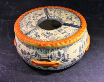 Könnte beinhalten: Ein weißer Keramik-Teekannenwärmer mit blau-weißem Blumenmuster und orangefarbenem Rand. Der Wärmer hat ein Loch in der Mitte für eine Teekanne und zwei Griffe an den Seiten.
