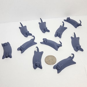 Op de afbeelding: Een verzameling van negen kleine, donkergrijze, 3D-geprinte kattenbeeldjes in verschillende liggende houdingen, elk met een zichtbare gelaagde textuur. Een Amerikaanse kwartmunt ligt tussen de beeldjes als referentie voor de grootte.