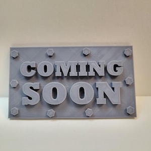 May include: A gray rectangular sign with the words "COMING SOON" in large, raised letters. The sign is adorned with hexagonal shapes around the edges, giving it a 3D printed appearance.