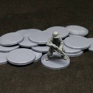 Pode incluir: Uma pilha de 12 bases de plástico cinzas para miniaturas de jogos de tabuleiro. As bases têm um diâmetro de 25 mm e são perfeitas para jogos de guerra, jogos de RPG e outros jogos de tabuleiro. Um soldado miniatura de plástico cinza está em pé sobre uma das bases.