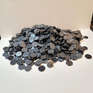 May include: A large pile of black, circular gaming bases with various textured surfaces. The bases are made of a dark material and are scattered across a white surface. A few bases are separated from the main pile.